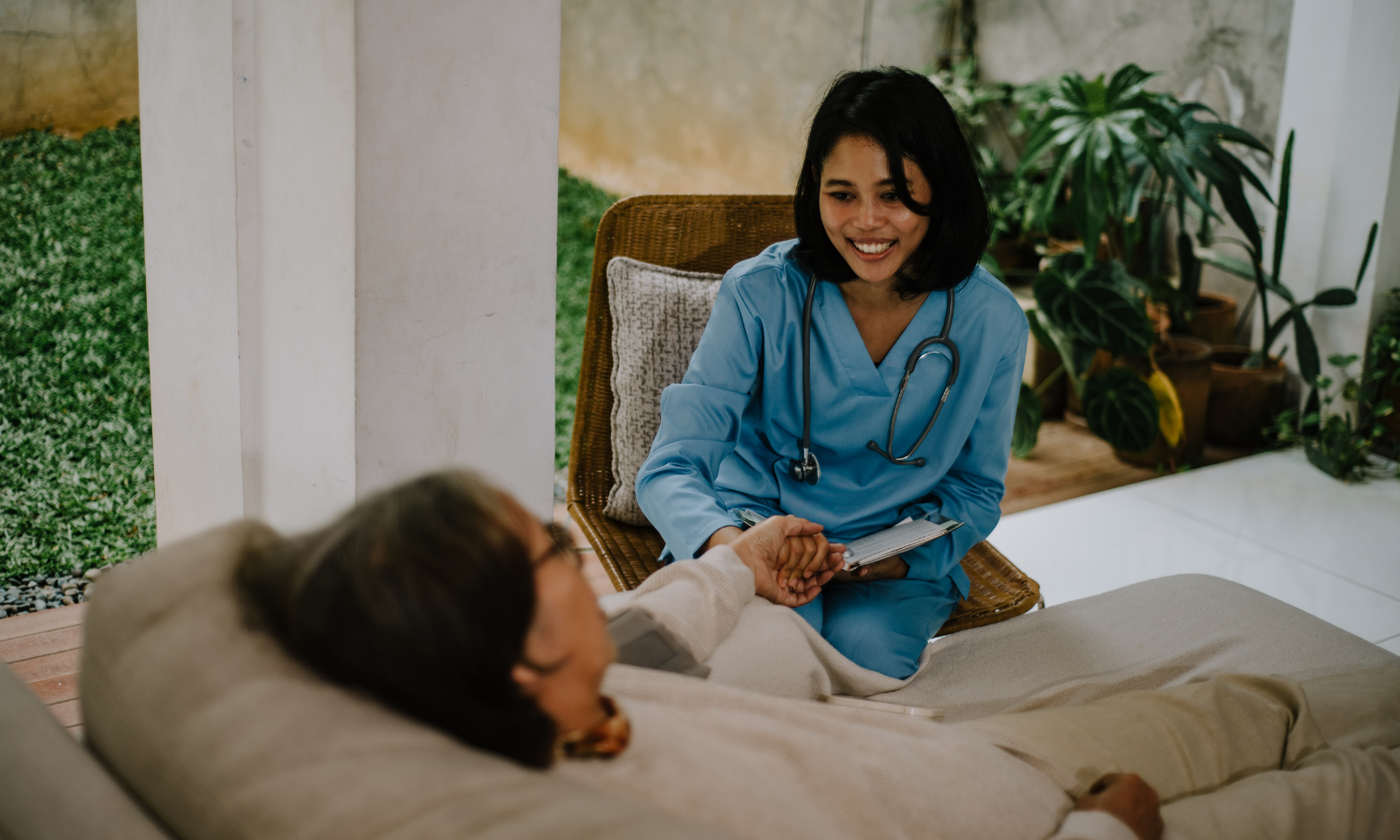 A licensed healthcare provider in scrubs with a stethoscope visiting a patient at home, sitting together in a calm, sunlit setting